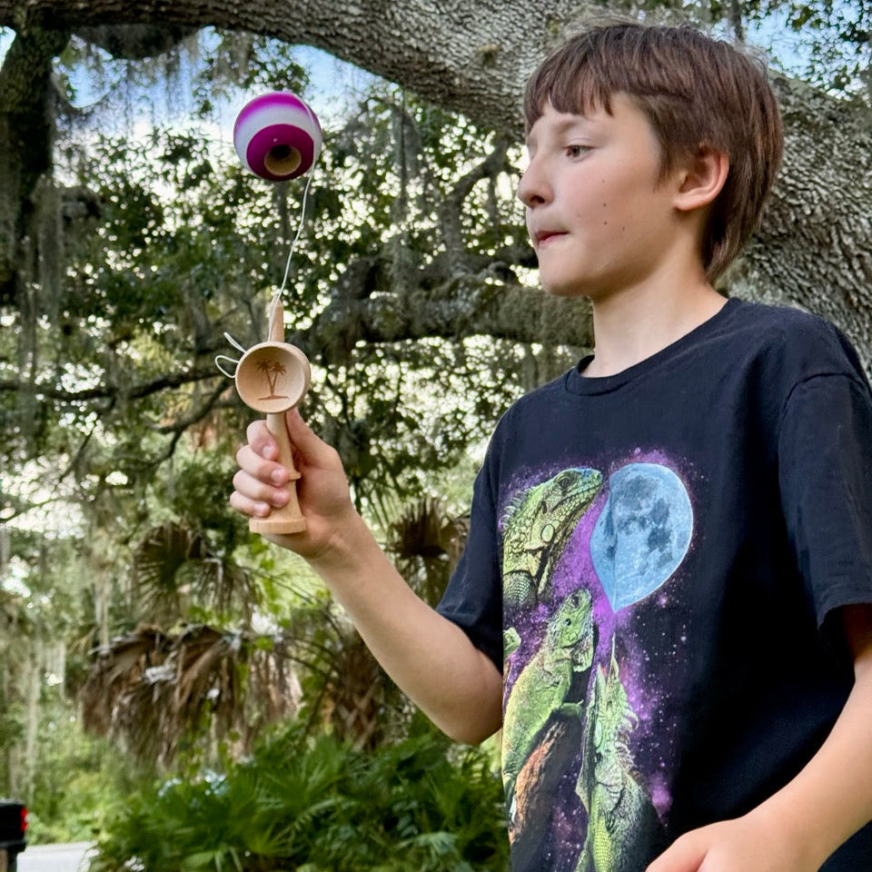 Boy wearing a black shirt, standing in a field, playing with a BK V2 Rubber Kendama that's purple and white. 
