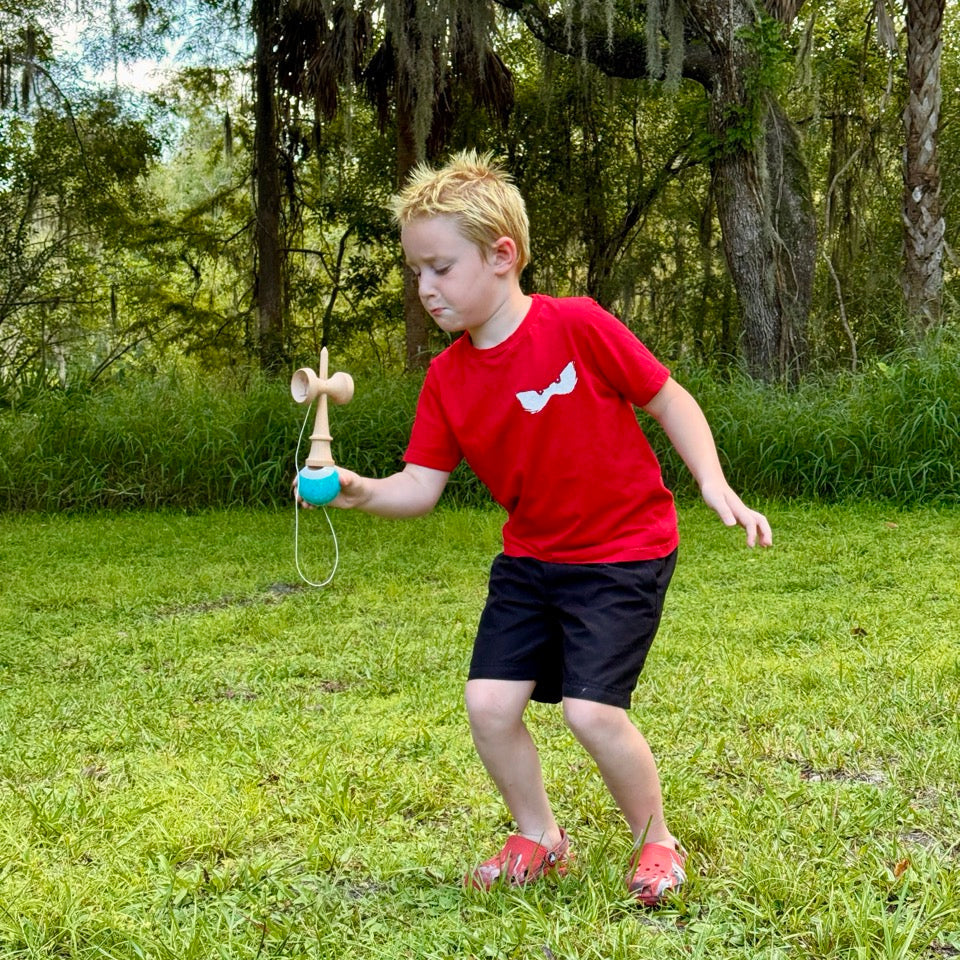 Boy wearing a red shirt, playing kendama in a field. 